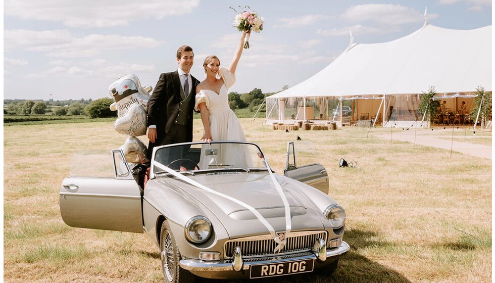 The Ultimate Wedding Planning Checklist | Just a married bride and groom are standing in their vintage car and celebrating their wedding in front of their wedding marquee.