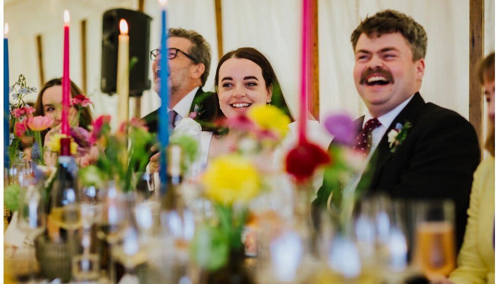 The Wedding Present Company | Smiling bride and groom behind the wedding table with spring-inspired floral decor and colourful candles.