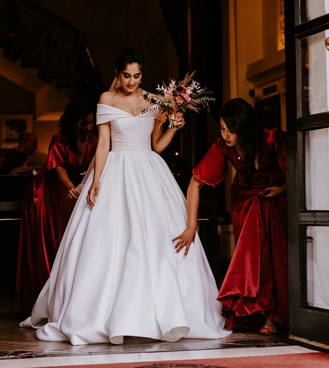 Bride leaves church holding a beautiful autumn wedding flower, while her bridesmaids fixes bride's dress. 