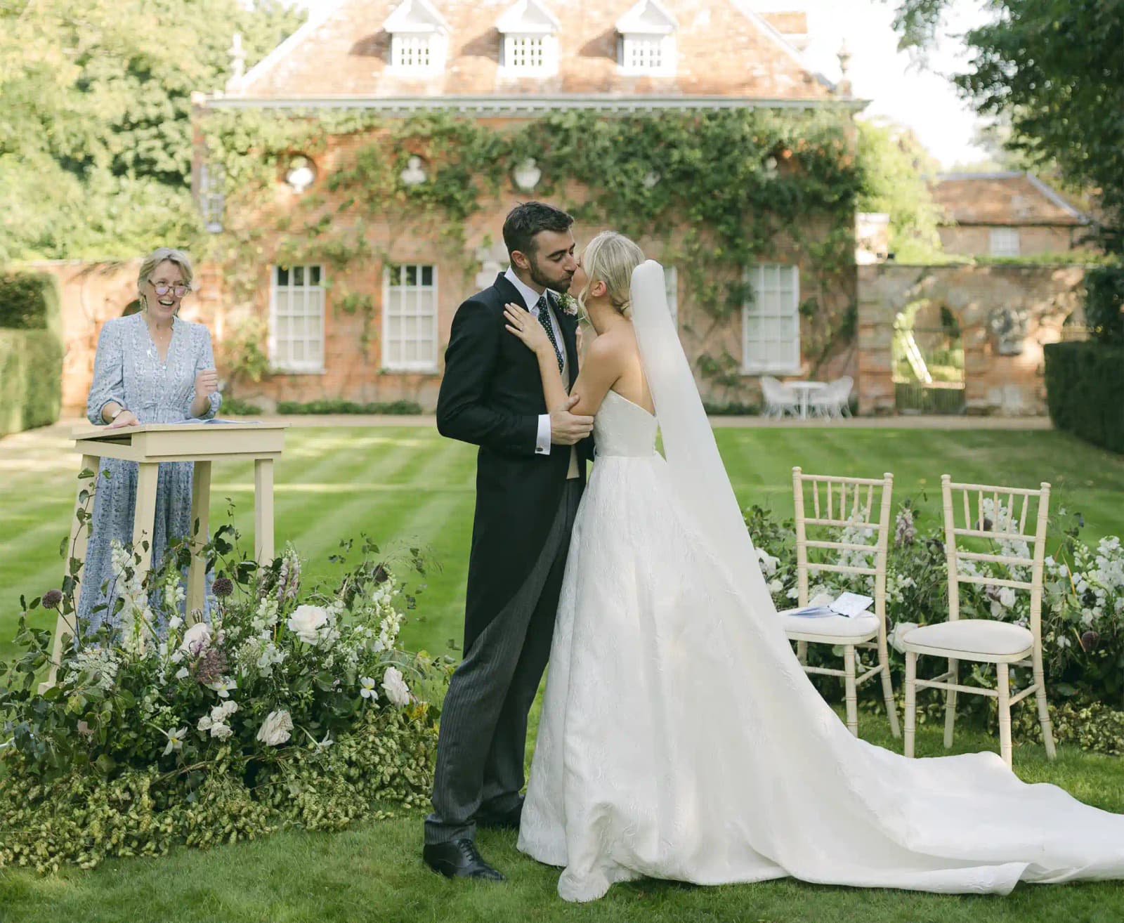  A bride and groom share a kiss during their outdoor wedding ceremony with officiant in front of ivy-covered brick house.
