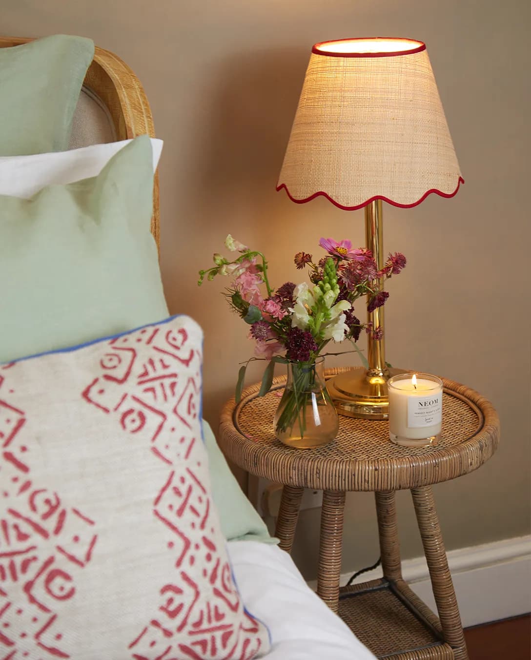 A rattan bedside table next to bed with patterned cushions features wedding presents including a scalloped lampshade from Matilda Goad, a bud vase with flower.
