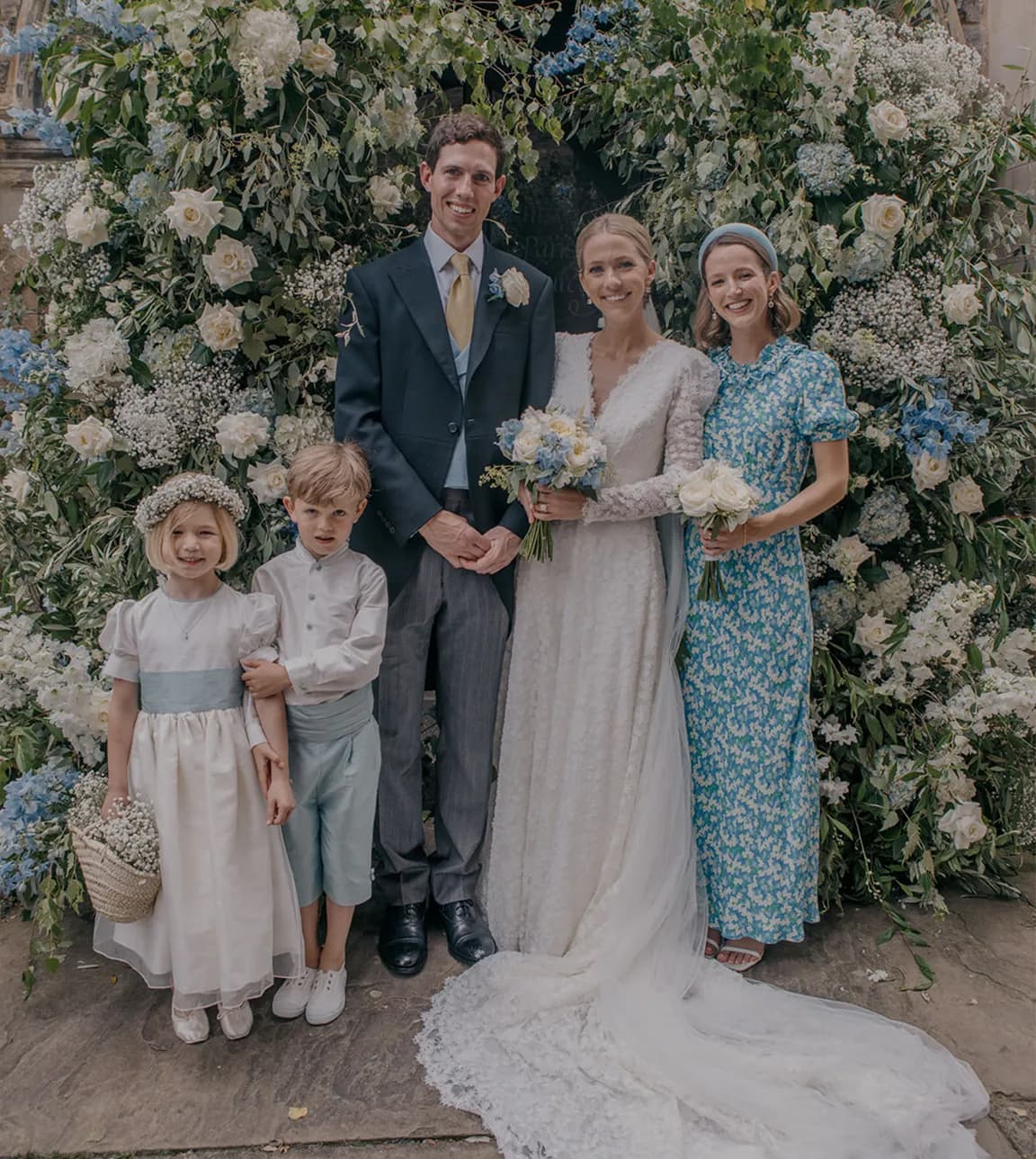 Traditional English bride and groom pose with page boy, flower girl and the maid of honour next to a floral arch.