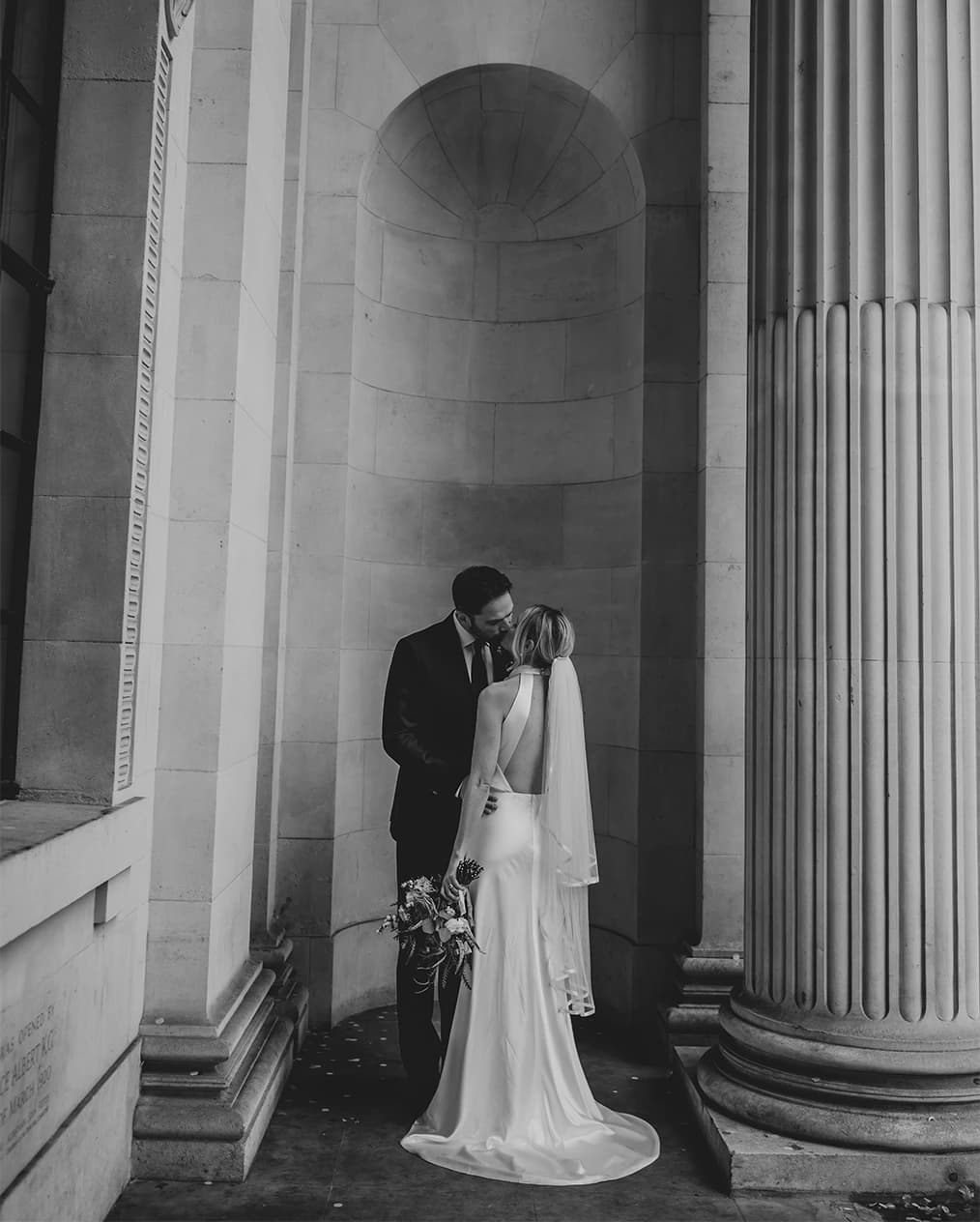 Husband and wife intimately kissing next to a large stone pillar
