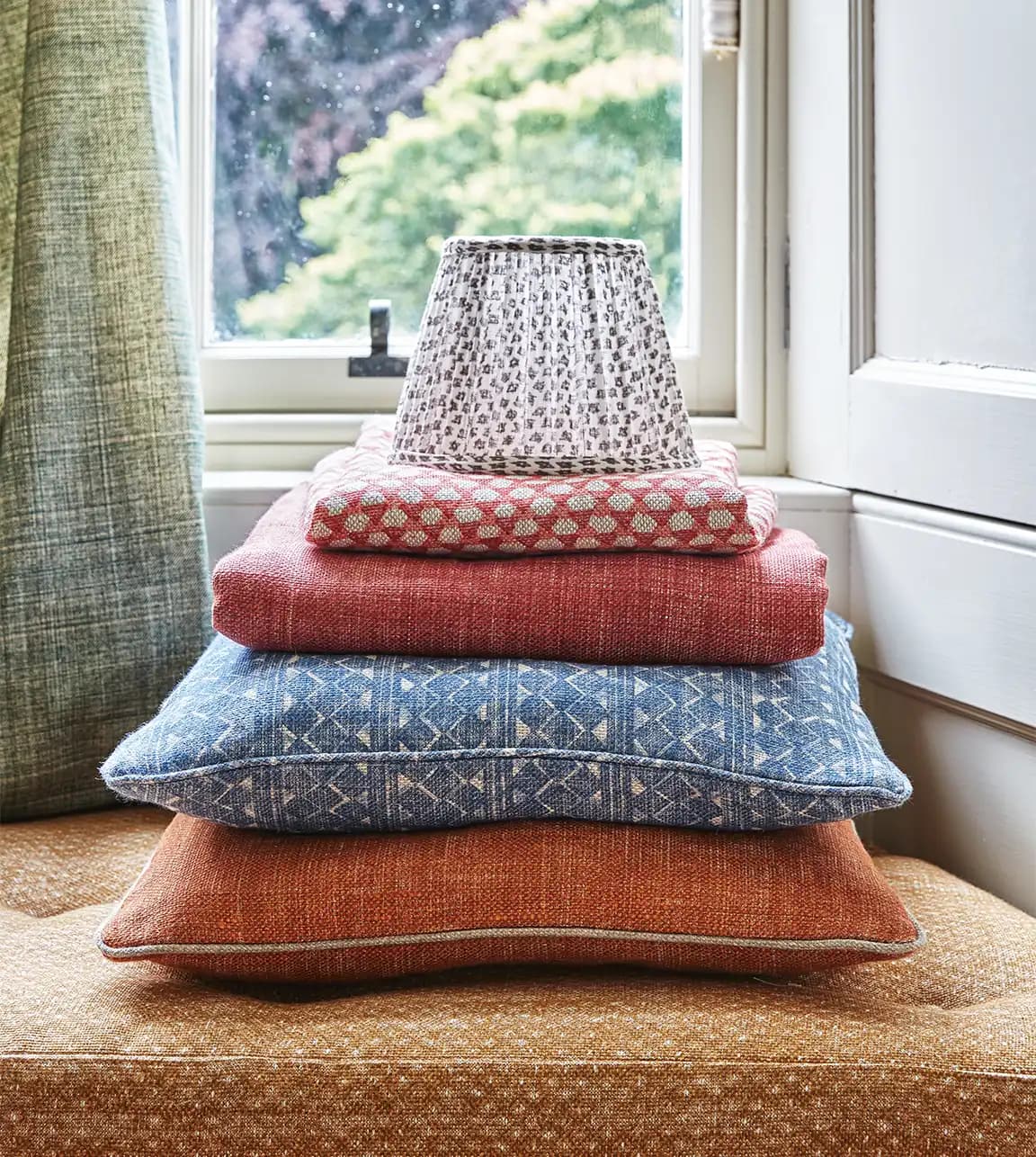 A pile of Fermoie patterned lampshade and cushions is styled on a bay window bench at home.
