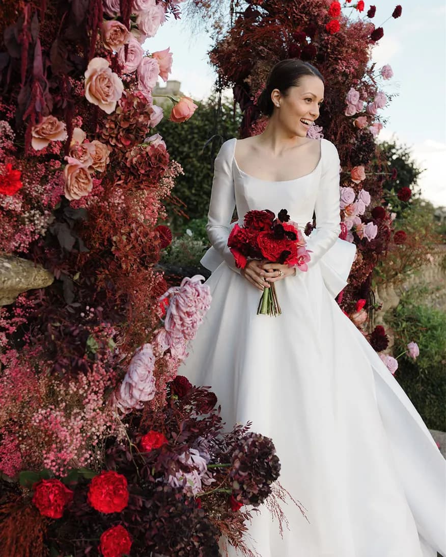 Smiling bride in demure wedding dress stands next to burgundy floral decor arch