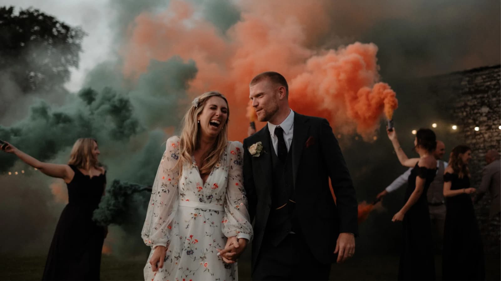 Bride and groom laughing while bridesmaids set off smoke flares at their festival-themed Cornwall wedding