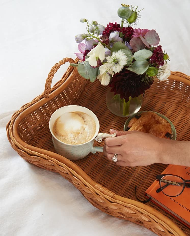 A female hand with engagement ring takes a mug with coffee from a stylish rattan tray.