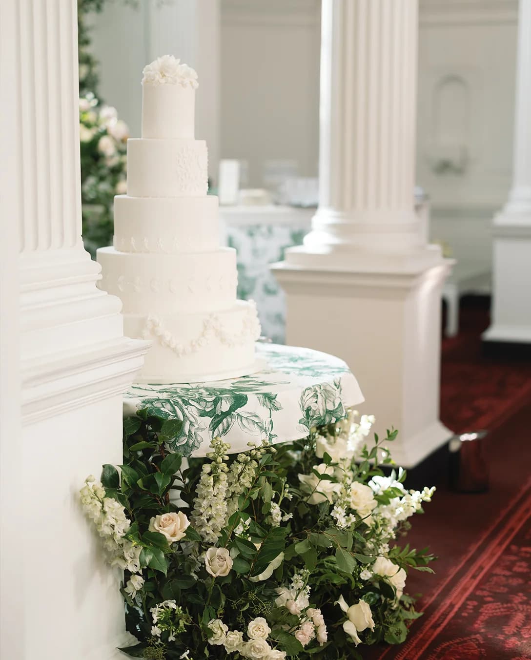 Five tier wedding cake with piping is placed on a table decorated with white and green florals.