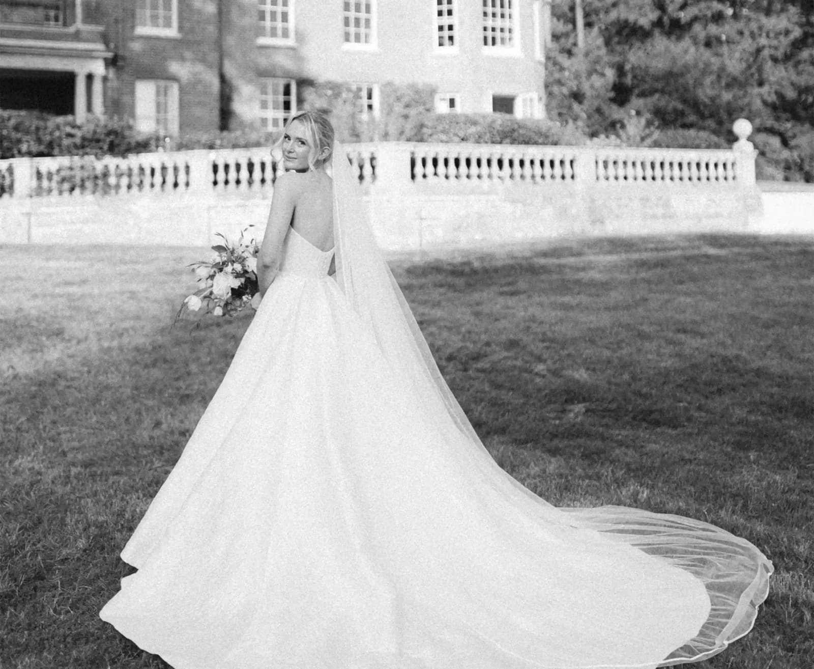 A beautiful bride in Sassi Holford strapless dress and veils stands in front of Georgian manor with railing.