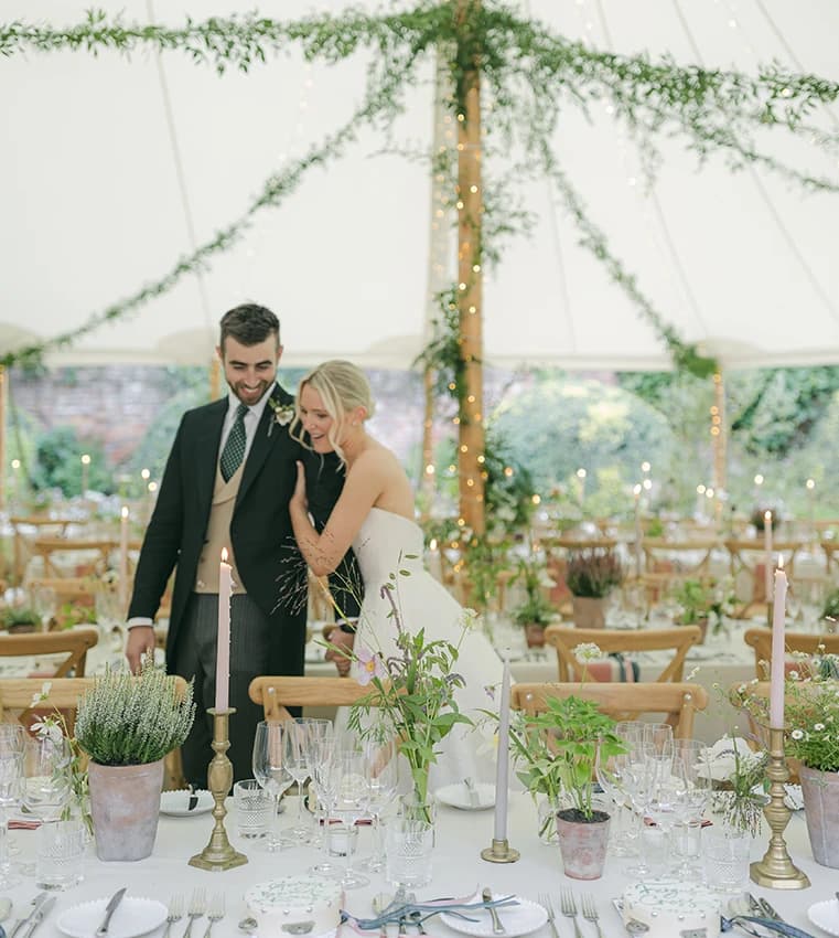 A bride and groom stand closely together, smiling and looking down at a beautifully set table in a traditional wedding marquee in Hampshire.