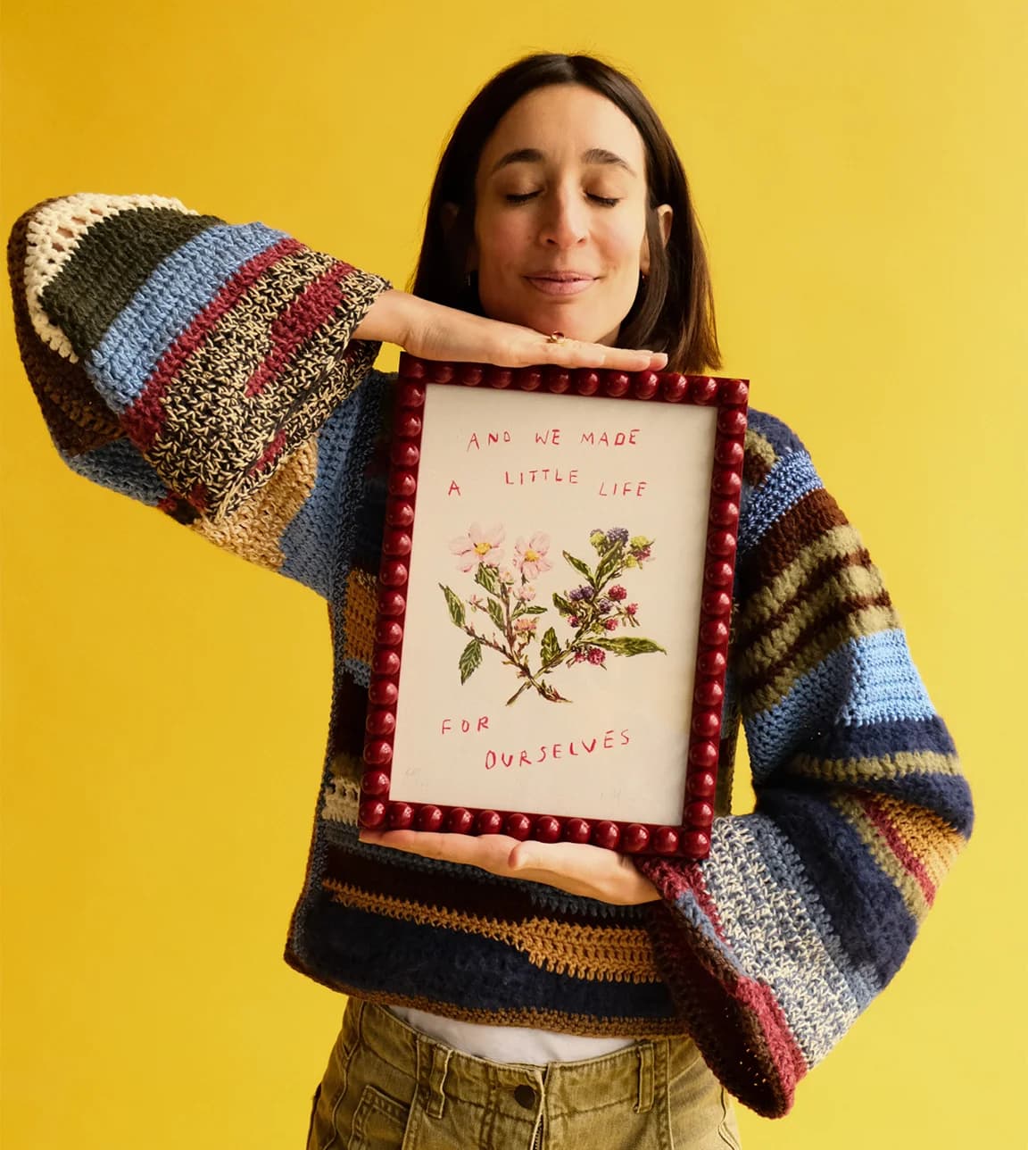 Laura Jackson stood against a yellow background wearing a multi-coloured, striped, knitted jumper. She's holding a maroon picture frame containing a print that says "And we made a little life for ourselves."