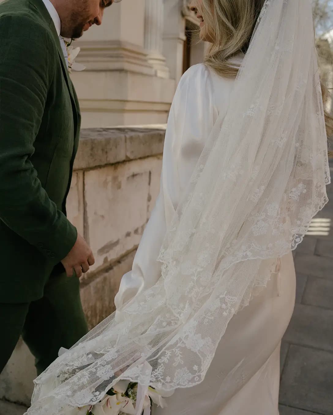 Groom in green linen suit and bride in silk wedding dress with vintage veil walk through the streets of London