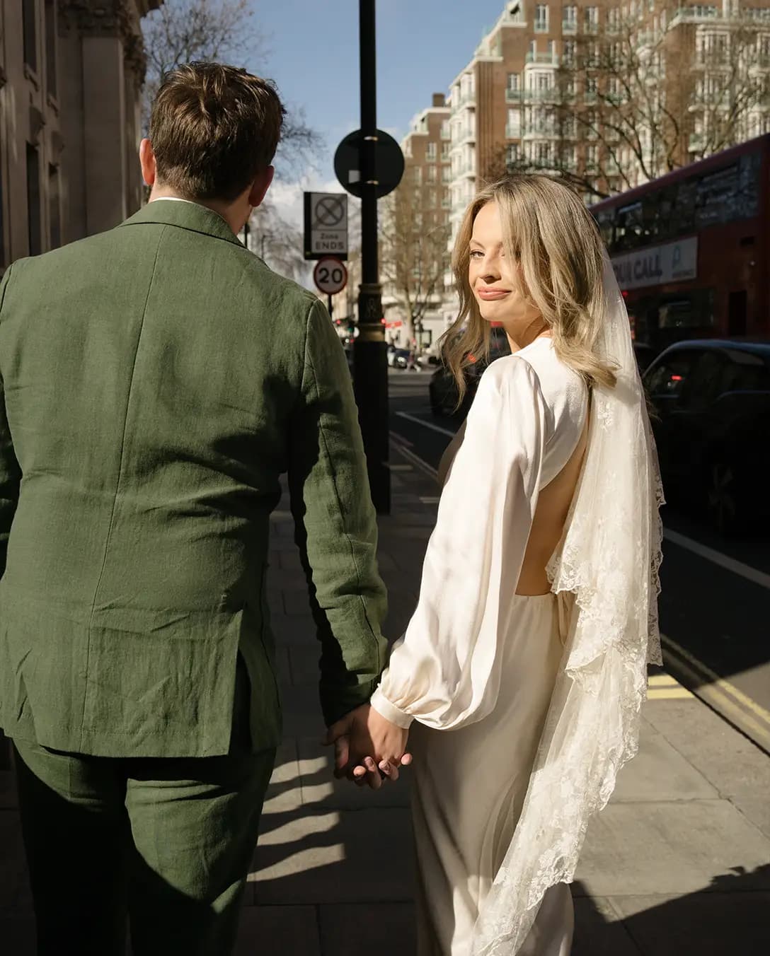 Groom in green linen suit and bride in silk wedding dress with vintage veil walk through the streets of London