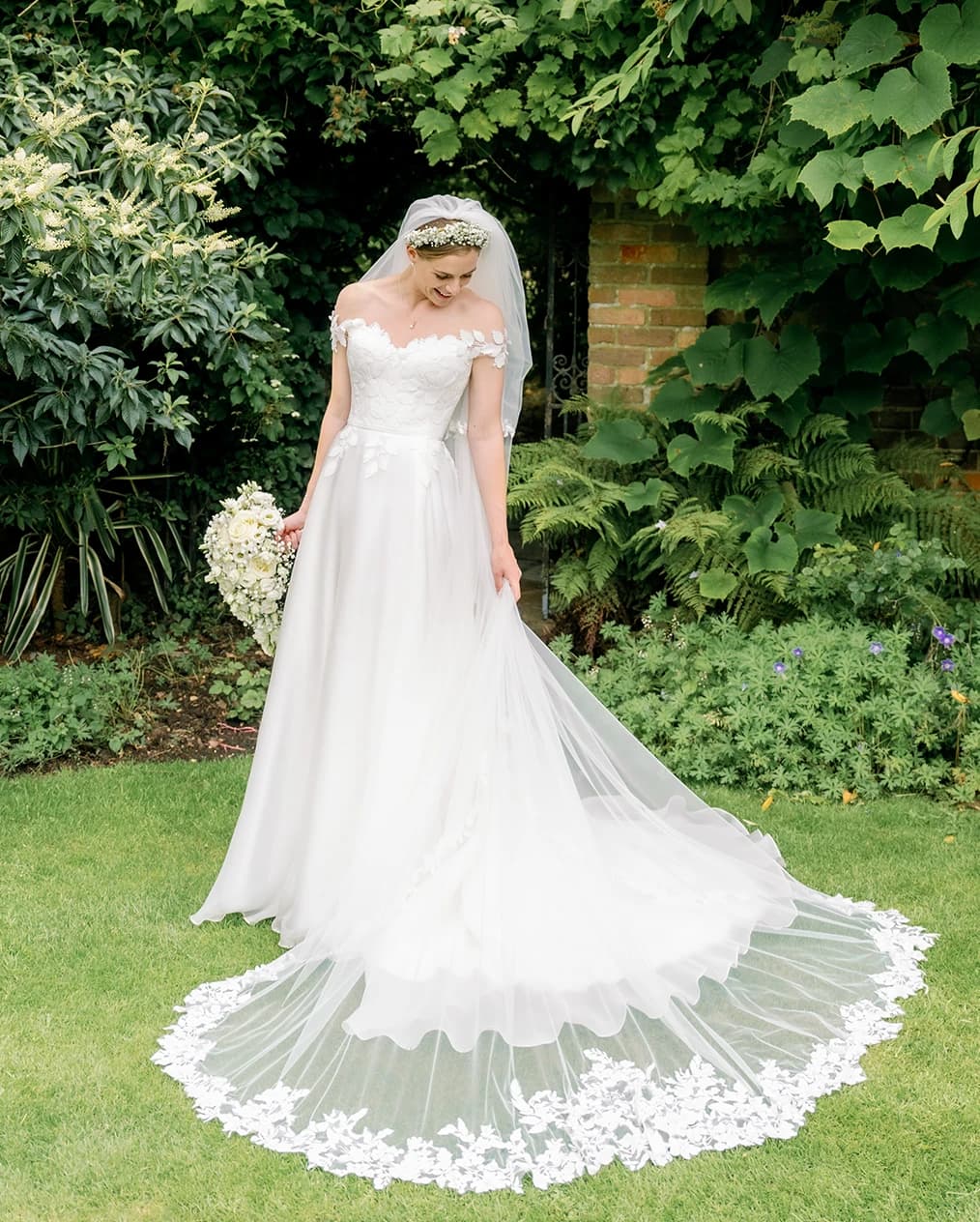 Bride wearing Suzanne Neville dress and long veil stands in the garden