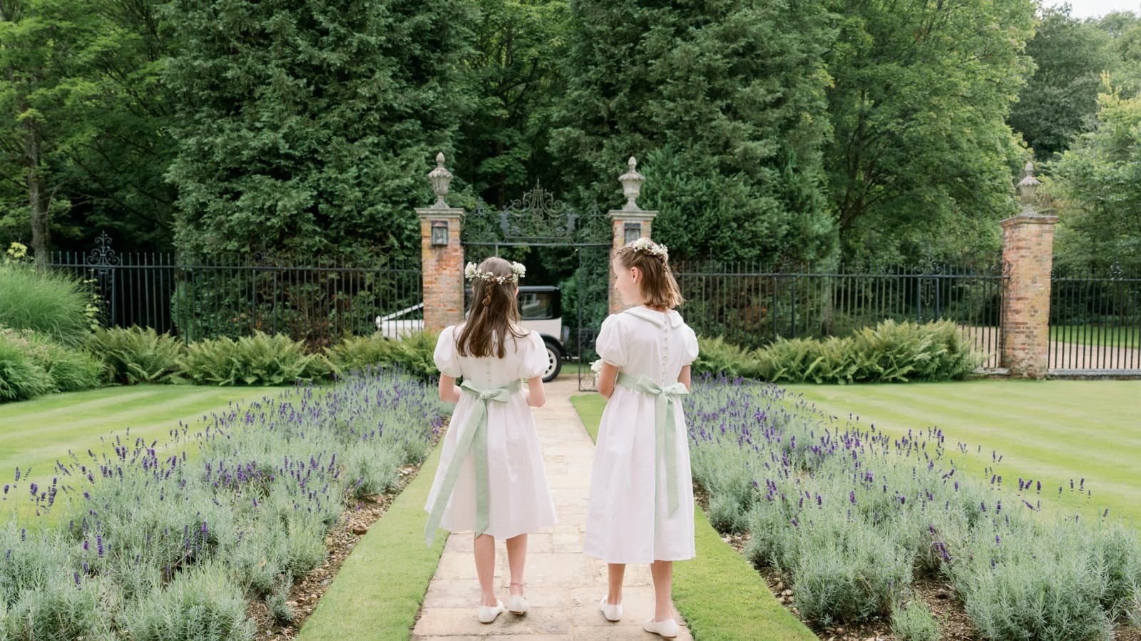 Flower girls wearing white dresses with pastel green bow walk through the garden