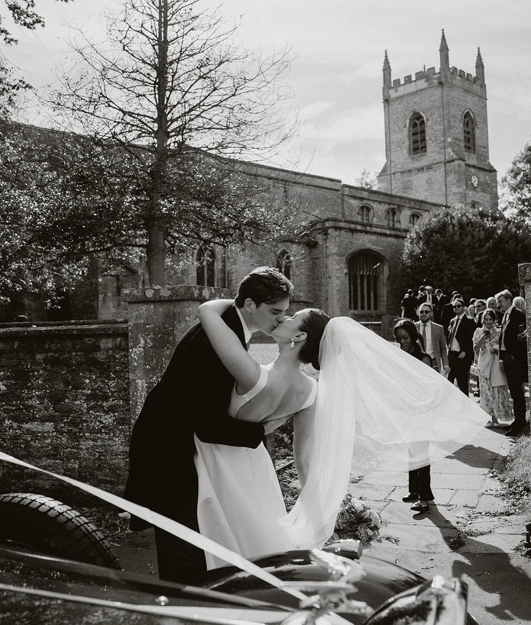Black and white photograph of the groom dipping the bride for a kiss beside the vintage car