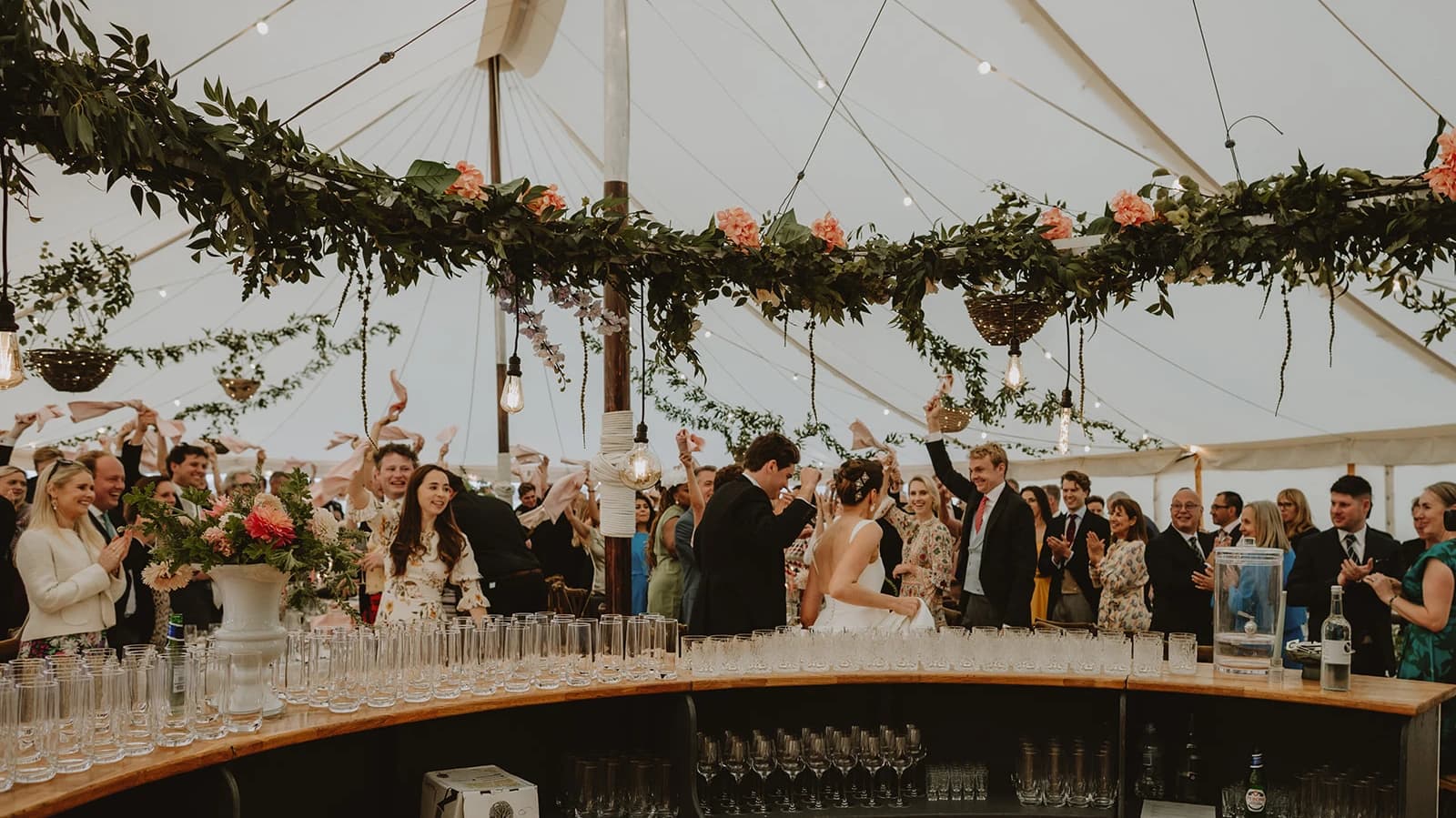 Bride and groom entering past the round bar as guests cheer beneath the garlands