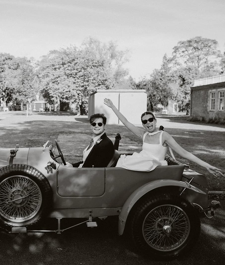 Black and white photograph of the couple waving from a vintage Bentley at Bicester Motion