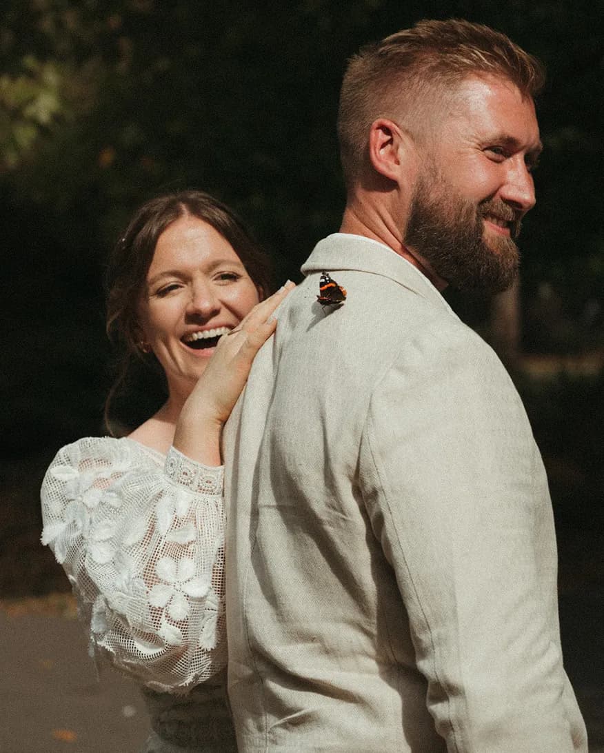 Bride in lace wedding dress looks at the butterfly that landed on groom's shoulder