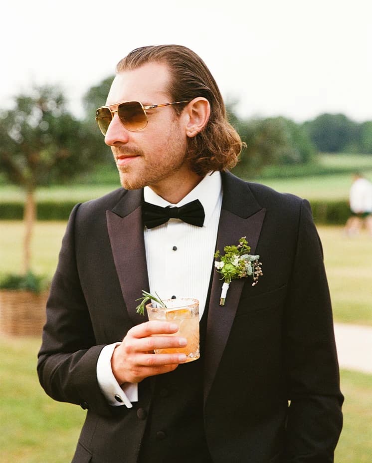 groom standing with a cocktail in hand wearing a bow tie and sunglasses