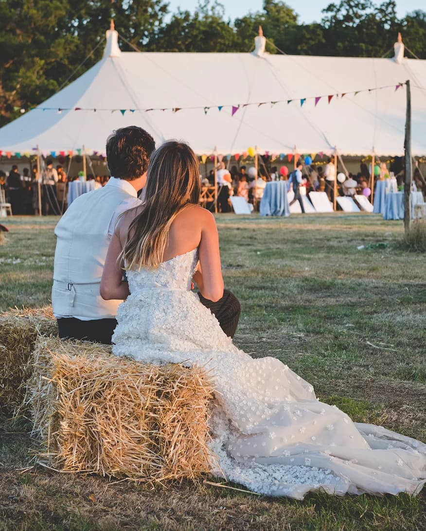 A bride and groom sit on a hay stack watching people outside their wedding marquee