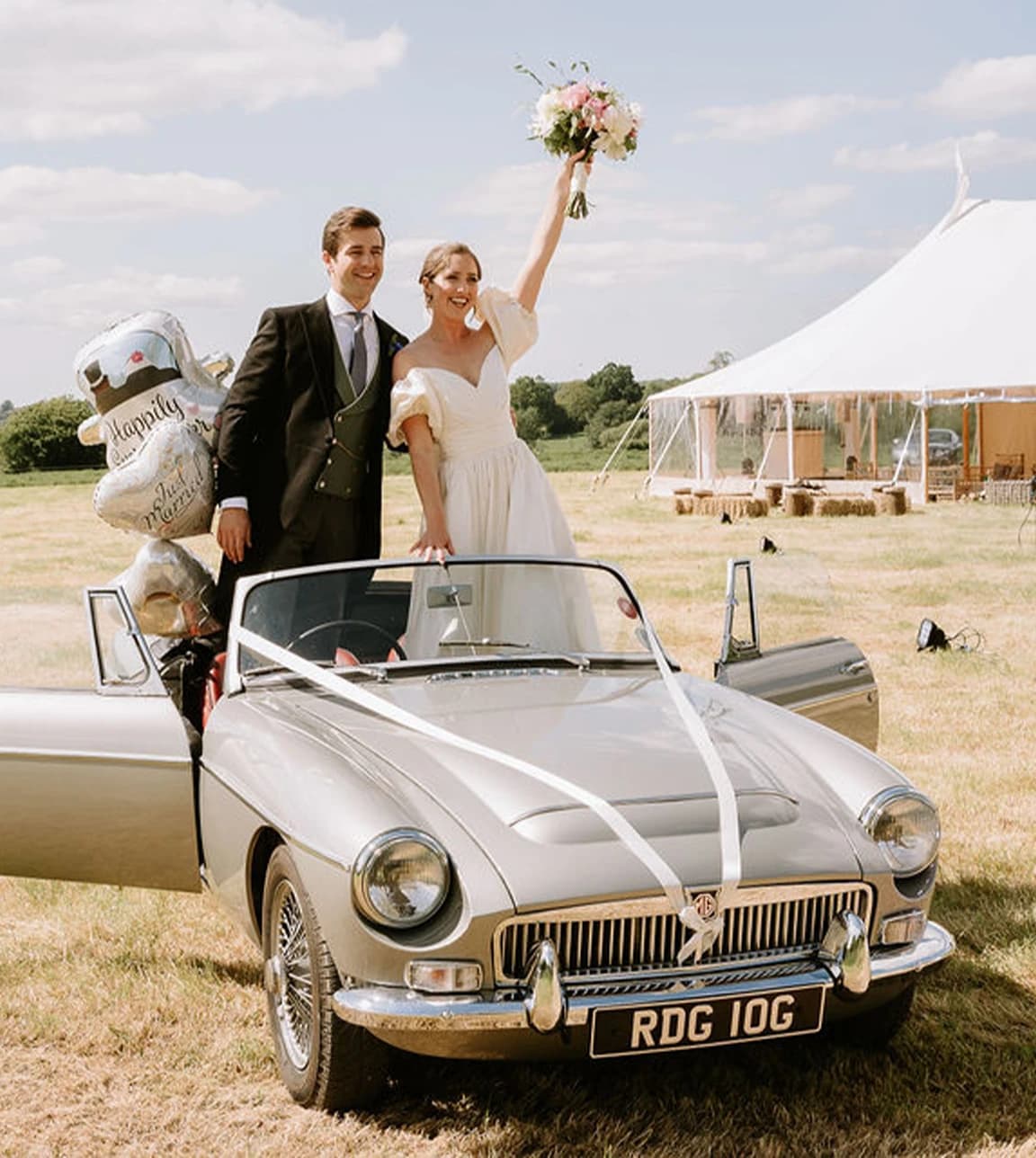 Bride and groom wave from a vintage car, standing next to traditional marquee