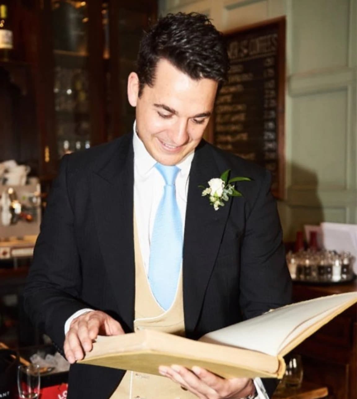 groom wearing morning dress featuring a blue tie and linen waistcoat looking down at a large book
