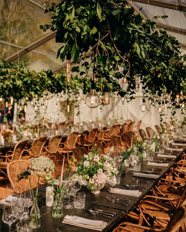 Wedding reception in a glass venue with wooden tables heavily decorated with foliage and pastel flowers