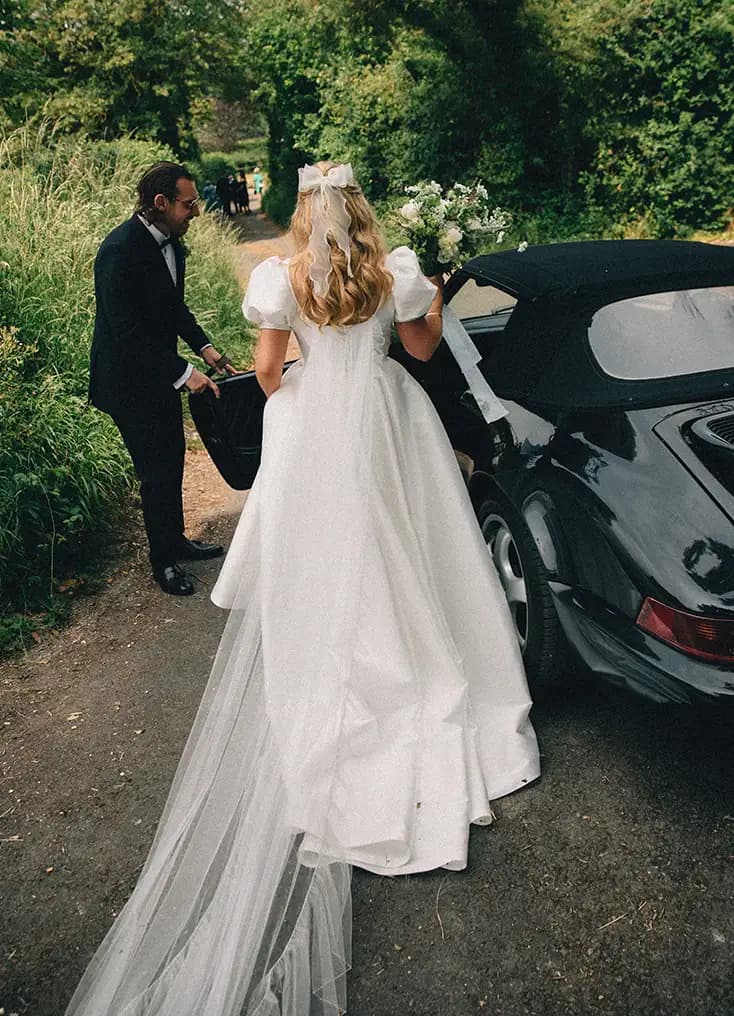 The Wedding Present Company bride with hair bow and groom gets into a black vintage car