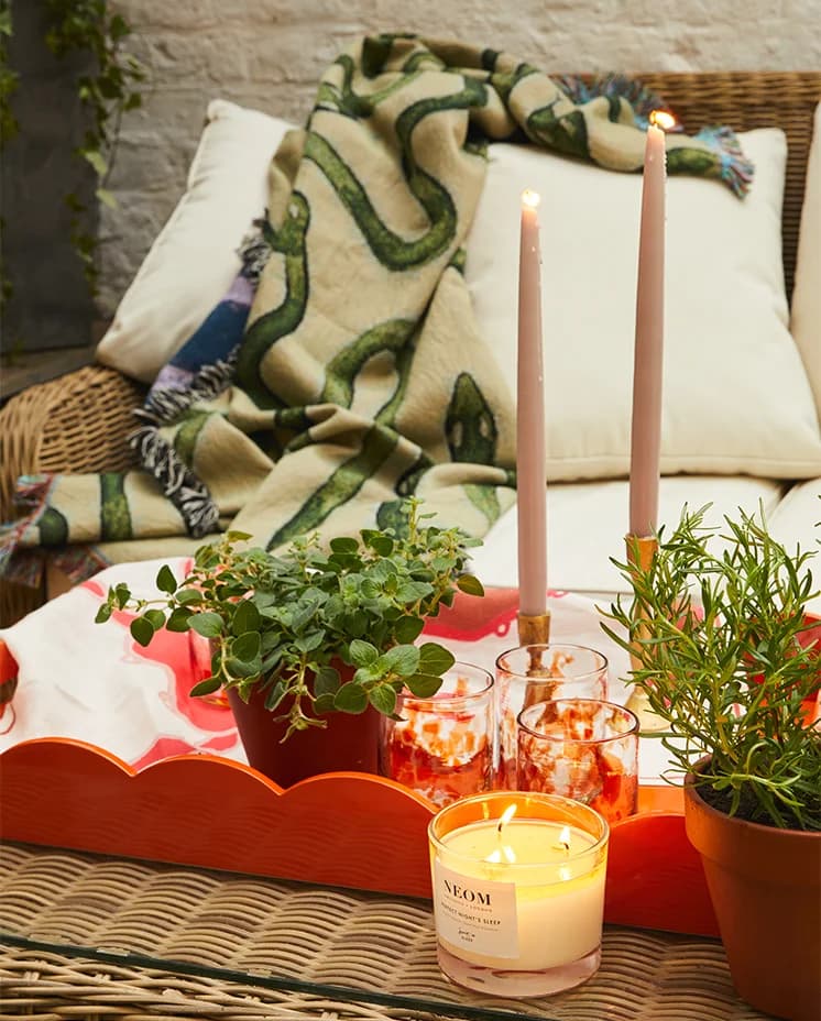 Orange scalloped Addison Ross Tray with candles, terrazzo tumblers and plants is placed on rattan furniture of cosy terrace.