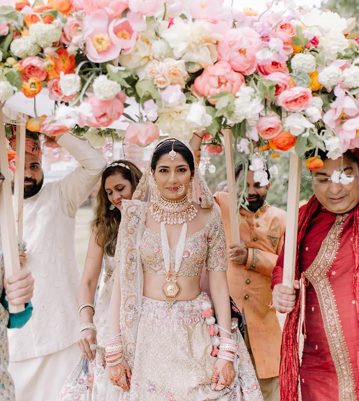 Bride walks through Indian bridal entry canopy from flowers, held by her family