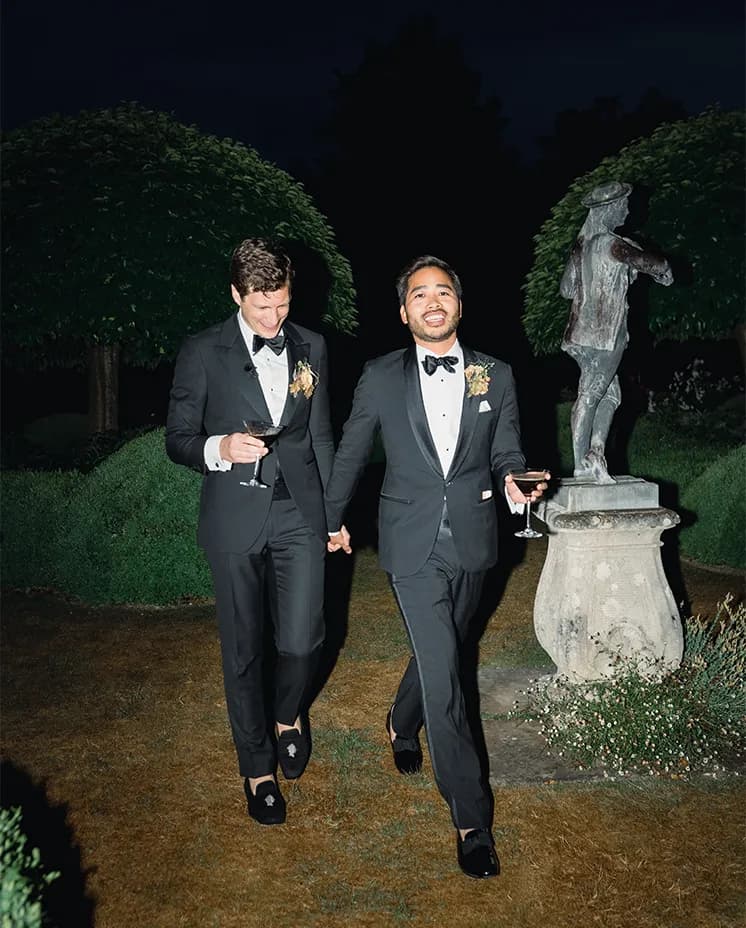 Two grooms in tux are walking together while holding hands and sipping on cocktails