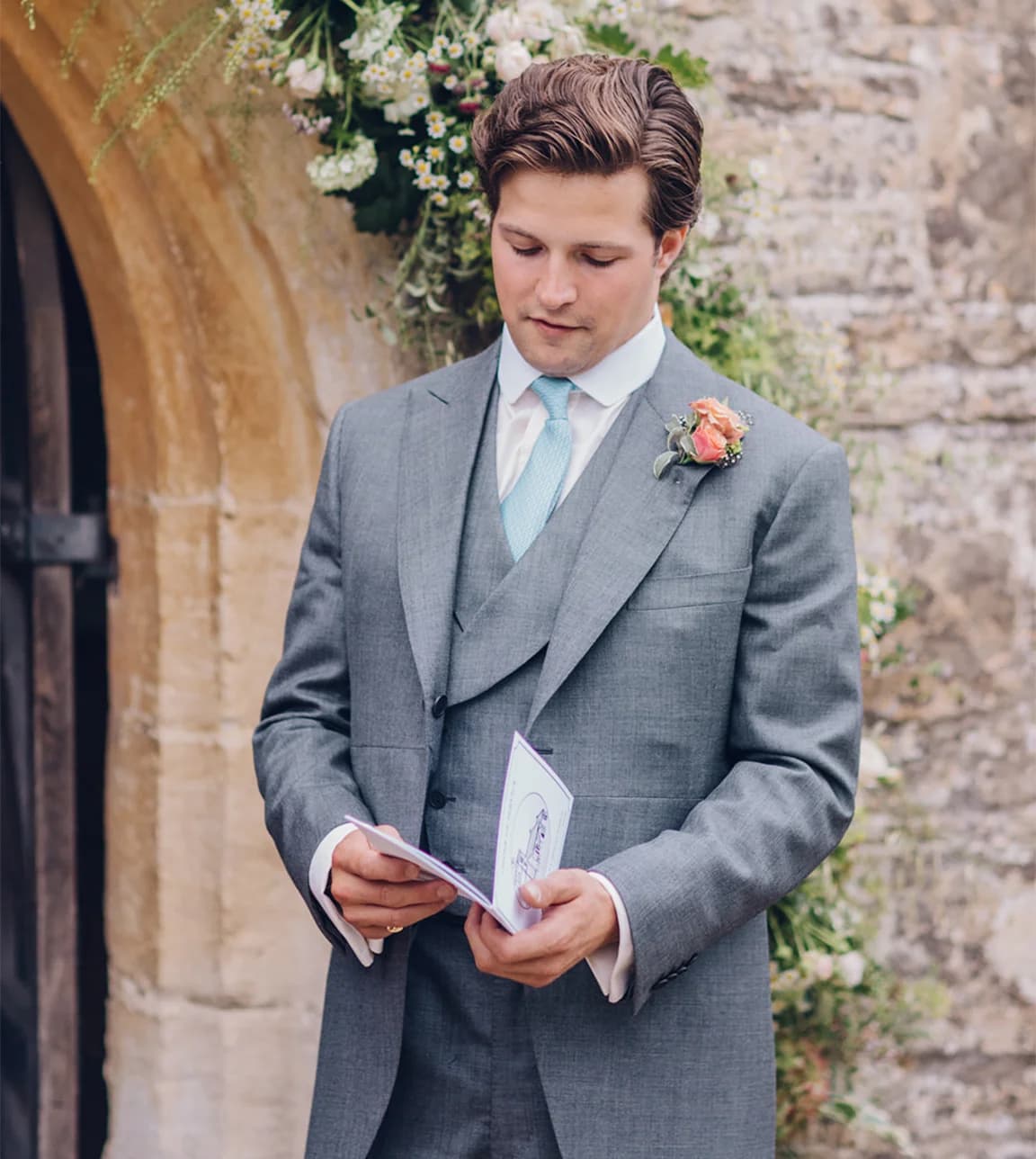 A groom in morning suit stands outside the church holding a wedding reading