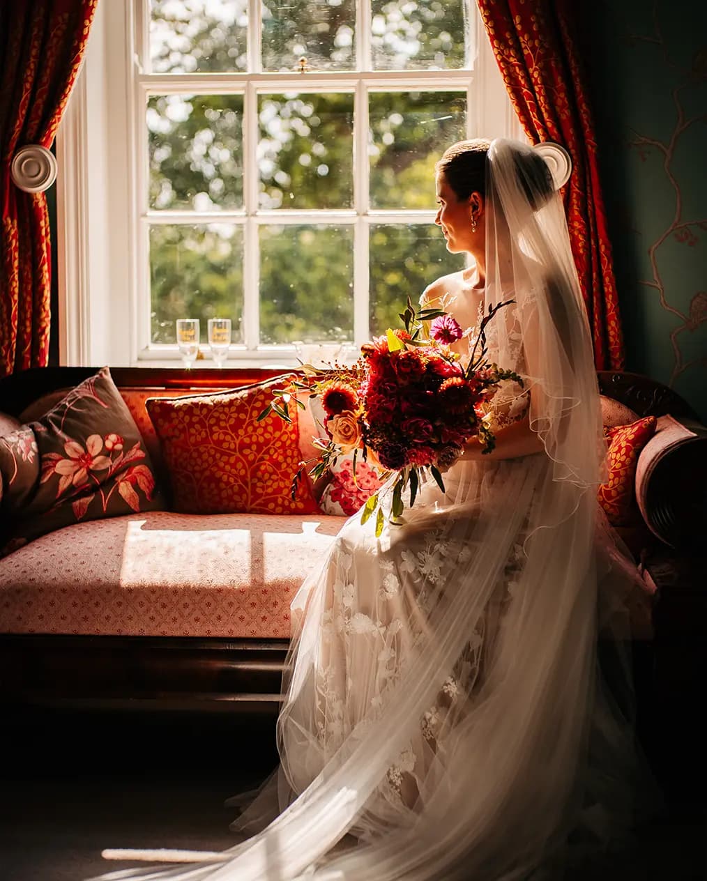 A bride sits in sunlit room on a chair holding autumn wedding flower bouquet