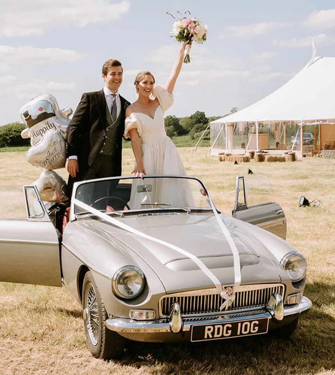 Bide and Groom stood up in the back of a vintage silver convertible car