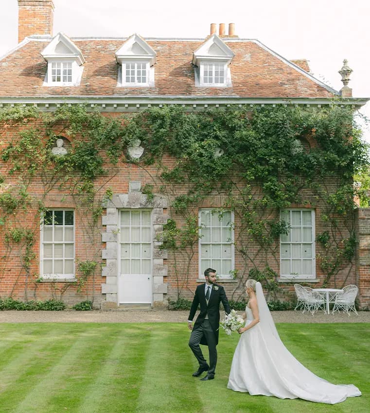 A bride and groom walk hand in hand in front of a charming manor house in Hampshire