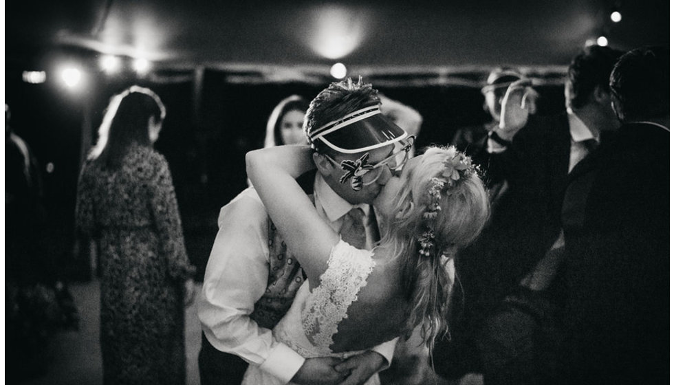 The bride and groom share a kiss on the dance floor wearing some party probs including sunglasses and a visor. 