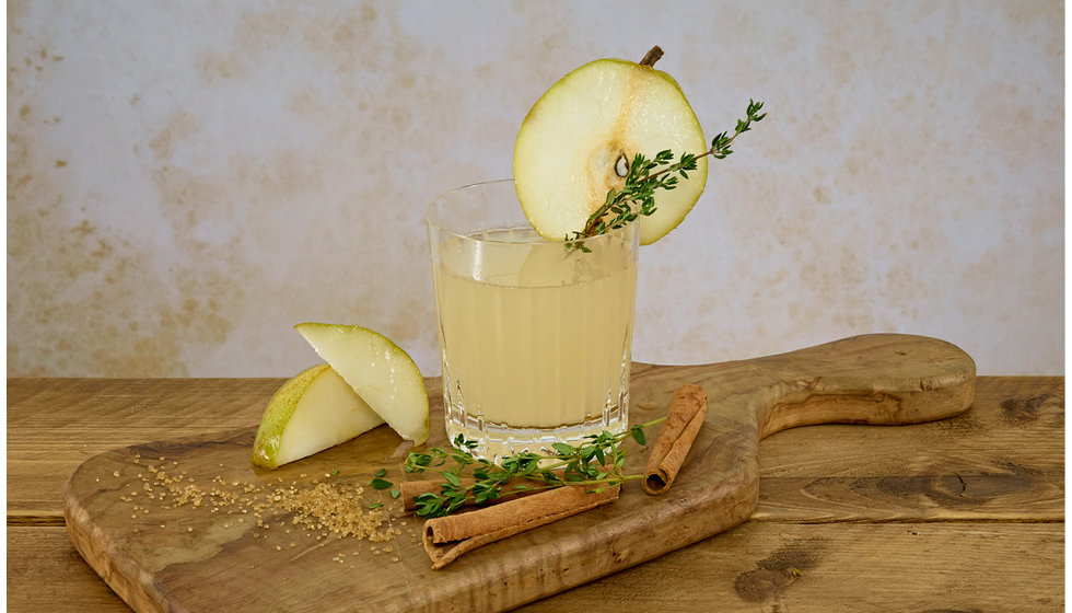 A spiced pear and thyme cocktail in a crystal tumbler on a wooden chopping board.