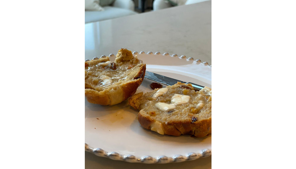 A hot cross bun cut open with butter melting on top on a white plate with a lovely pearl rim.