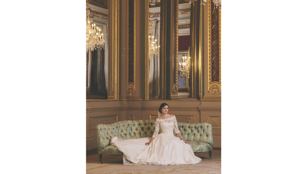 A bride on a sofa wearing a quarter sleeved wedding dress with a long train.