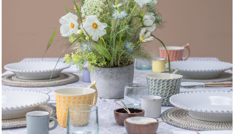 A spring breakfast table with spring flowers in the centrepiece. There is a printed tablecloth, colourful mugs, white beaded plates and little blue recycled glass tumblers. All using products from the Wedding Present Company's online showroom. 