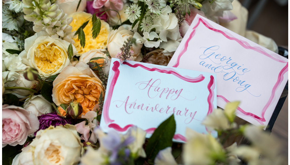Two illustrated cards saying 'Happy Anniversary' and one saying 'Georgie and Doug' on top of some beautiful roses.