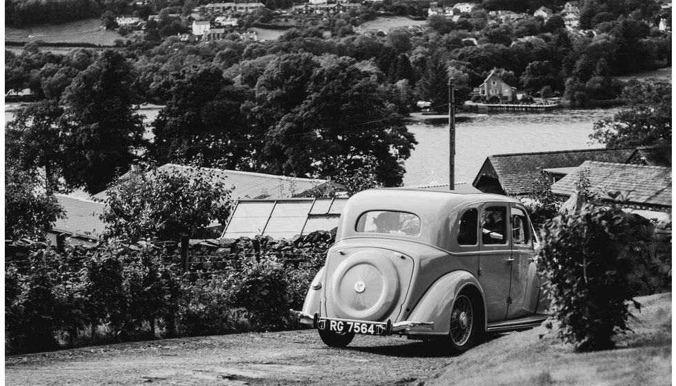 A black and white image of a vintage car driving down into a small village in the countryside.