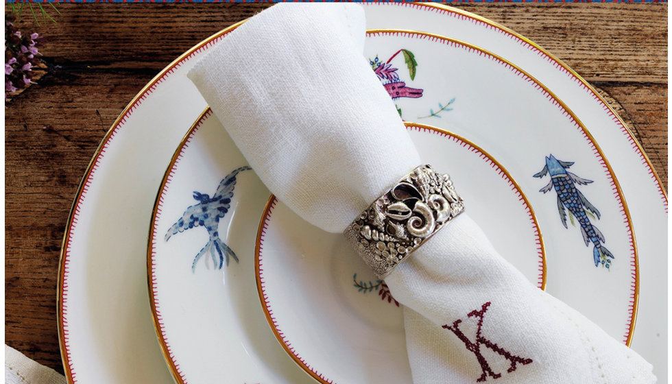 A stack of Kit Kemp Mythical Creatures plates with a embroidered napkin with a napkin ring.