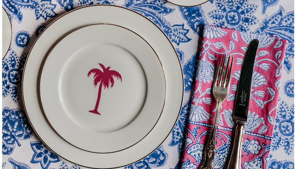 An Alice Peto pink palm tree side plate on top of a gold rimmed dinner plate on top of a blue and white floral tablecloth with a pink napkin and silver cutlery.