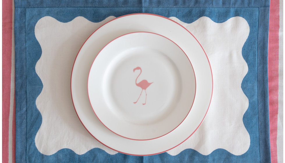 An Alice Peto pink side plate with a Flamingo on it, on top of a pink rimmed dinner plate on a blue scalloped fabric placemat.