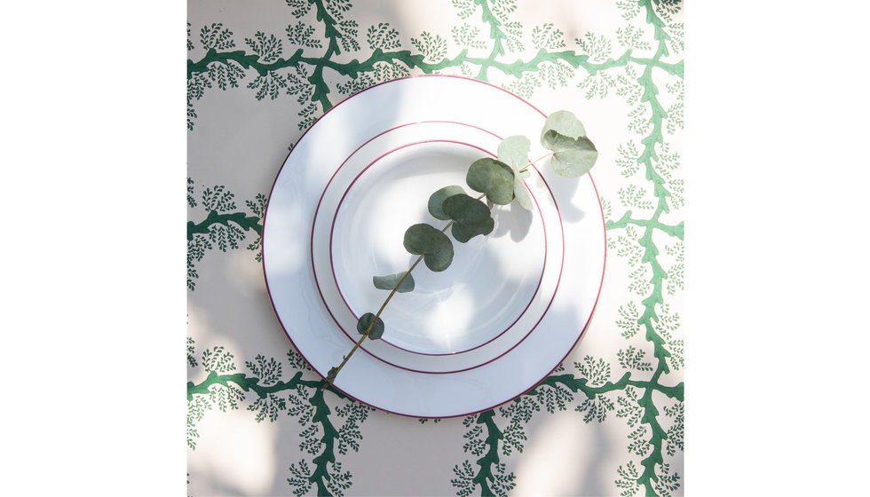 A dinner plate, starter plate and bowl rimmed with cerise with a stem of eucalyptus on it on a green and white tablecloth. 