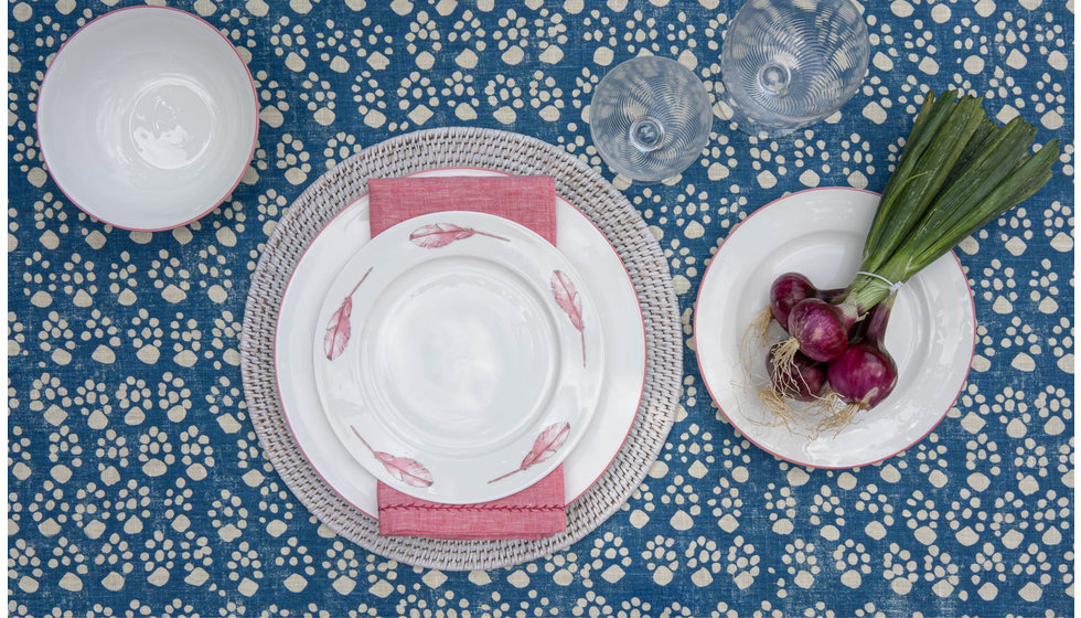 A table laid with a blue print tablecloth, a white rattan placemat with a pink dinner table, a pink side platw with feathers and a pink napkin.