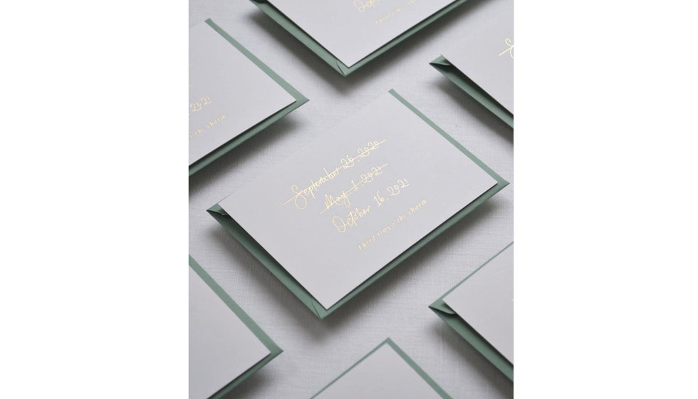 Wedding invitations with the two previous planned dates crossed out with the new wedding day in gold foil on green card.