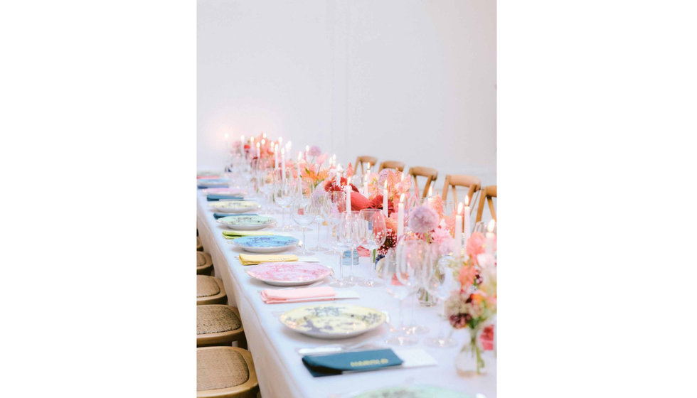 A long table lined with multi coloured dinner plates, pink candles and pink dried flowers. 
